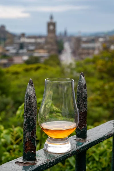 Glass of single malt scotch whisky and view from Calton hill to park and old parts of Edinburgh city in rainy summer day, Scotland, UK