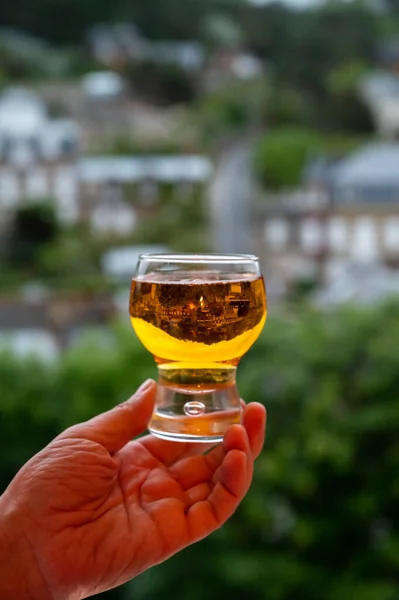 Hand holding one glass of apple cider drink and houses of Etretat village on background, Normandy, France