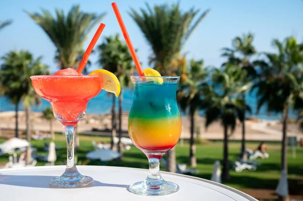 Colourful cold Strawberry daiquiri and Rainbow Paradise cocktails drinks served in glasses at outdoor cafe overlooking blue sea, palm trees, relax and holidays at sea