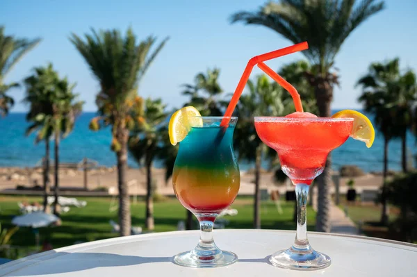 Colourful cold Rainbow Paradise and Strawberry daiquiri cocktails drinks served in glasses at pool bar overlooking blue pool, sea and palm trees, relax and holidays at sea