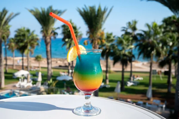 Colourful frozen Rainbow Paradise cocktail drink served in glass at pool bar overlooking blue pool, sea and palm trees, relax and holidays at sea