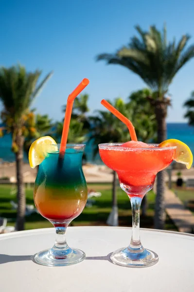 Colourful cold Rainbow Paradise and Strawberry daiquiri cocktails drinks served in glasses at pool bar overlooking blue pool, sea and palm trees, relax and holidays at sea