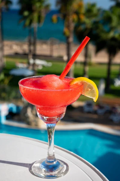 Colourful frozen pink Strawberry daiquiri cocktail drink served in glass at pool bar overlooking blue pool, sea and palm trees, relax and holidays at sea