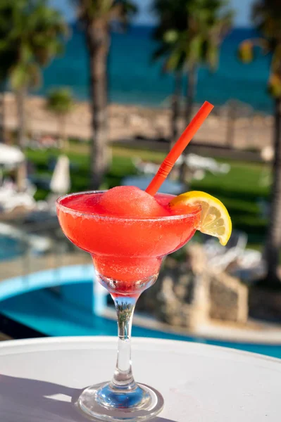 Colourful frozen pink Strawberry daiquiri cocktail drink served in glass at outdoor cafe overlooking blue pool, sea and palm trees, relax and holidays at sea