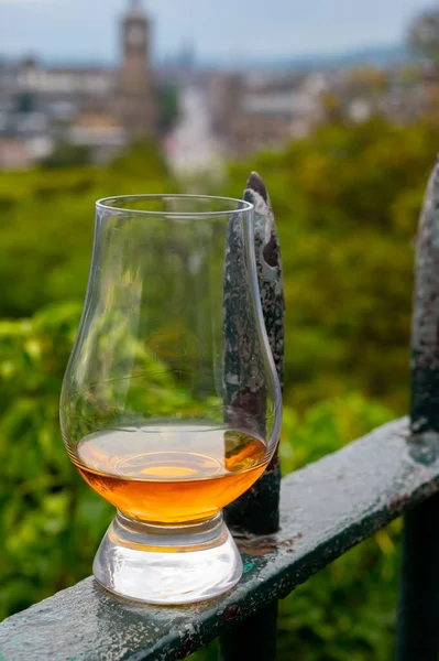 Glass of single malt scotch whisky and view from Calton hill to park and old parts of Edinburgh city in rainy summer day, Scotland, UK