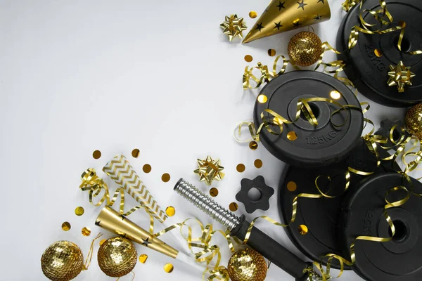 Heavy dumbbells weight plates, golden decorations, confetti, party hat, horn blowers for New Year\'s Eve celebration. Healthy fitness lifestyle flat lay concept. Gym, workout New Year\'s resolution composition with copy space on white background.