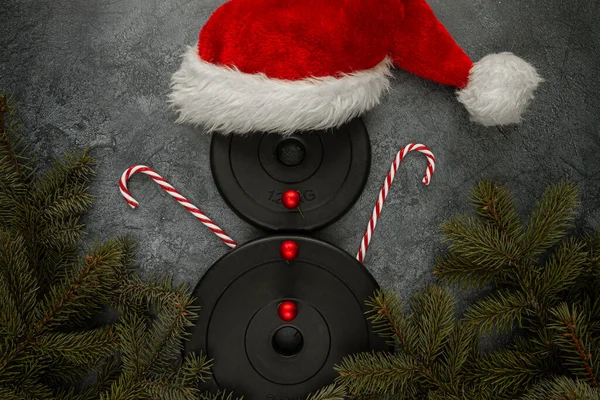 Heavy dumbbells weight plates, shaped as a snowman with red Santa Claus hat, candy canes as hands and decorative baubles as buttons. Healthy fitness lifestyle Christmas gym concept with tree branches.