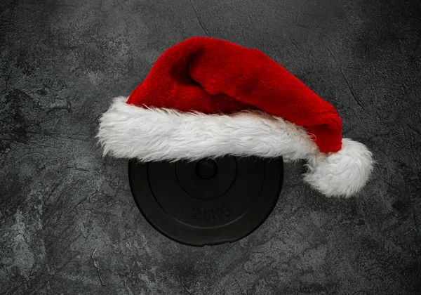 Heavy dumbbell barbell weight plate disc with red Santa Claus hat for Christmas. Healthy fitness gym lifestyle holiday season concept, flat lay composition.
