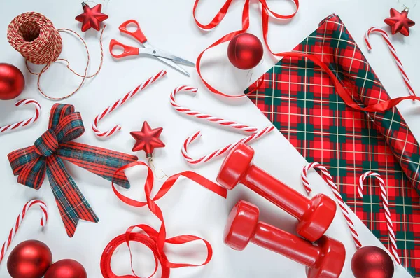 Gift wrapping paper and string, two red dumbbells, ribbon and bow, candy canes, scissors and decorative baubles. Exercise equipment as a Christmas gift idea. Healthy fitness lifestyle holiday season.