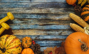Two yellow dumbbells with various pumpkins, squashes and gourds. Healthy gym fitness flat lay lifestyle autumn composition, with cones, corn on the cob and autumn leaves. Copy space on wooden background.