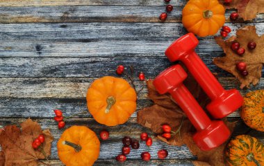 Two red dumbbells with pumpkins and autumn leaves. Healthy gym fitness lifestyle fall composition. Flat lay with copy space on wooden background, for Thanksgiving or Halloween.