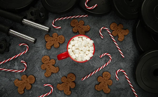 Heavy dumbbells barbell weight plates, gingerbread man cookies, candy canes and a mug of hot cocoa with marshmallows. Gym fitness lifestyle flat lay composition for Christmas. Healthy diet during holiday season concept.