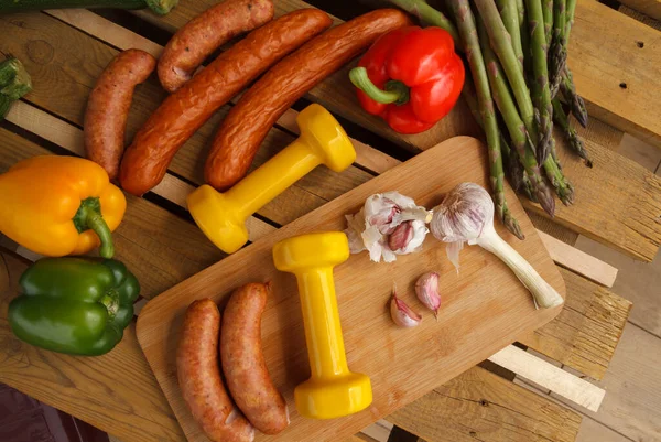Heavy dumbbells, vegetables and sausages for a grill. Fit barbecue party concept, fitness lifestyle and healthy dieting choice, gym workout composition. Cheat day temptation vs sticking to diet.