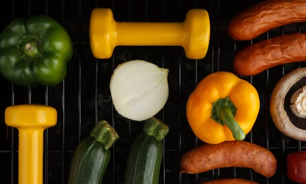 Heavy dumbbells with vegetables and sausages on a grill. Fit barbecue party concept, healthy fitness lifestyle, gym workout composition. Cheat day temptation vs sticking to diet.