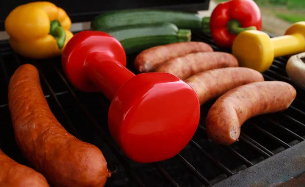Heavy dumbbells with sausages and vegetables on a grill. Fit barbecue party concept, healthy fitness lifestyle, gym workout composition. Cheat day temptation vs sticking to diet.