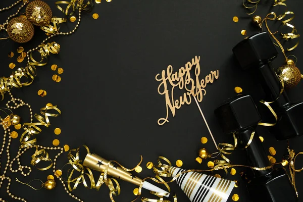 Black dumbbells, golden decorations, confetti, party hat and horn blower for New Year's Eve celebration. Healthy fitness lifestyle flat lay concept. Gym, workout New Year resolution composition, with copy space on black background.