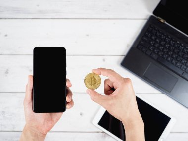 Elinizde altın Bitcoin ve cep telefonu, tahta masa arkasında dizüstü bilgisayar ve tablet var.