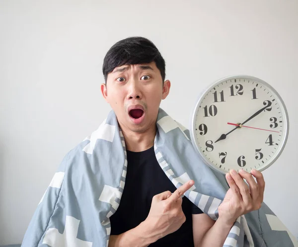 Asian man cover blanket point at clock in his hand with shocked face late concept white background