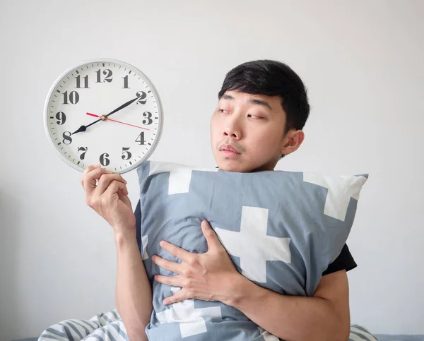Young man wake up hugging pillow and look at clock in hand feel bored at face on white isolated wake up late concept