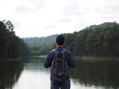 Gezgin konseptiyle göl ve dağ arasında duran Asyalı adam sırt çantası vadi ve nehir altyapısı olan çanta taşıyor.