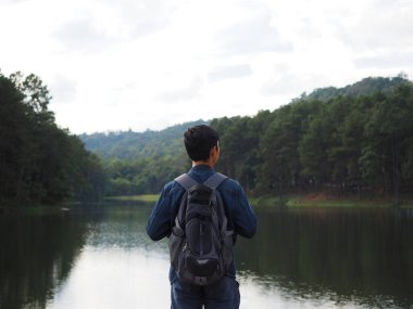 Gezgin konseptiyle göl ve dağ arasında duran Asyalı adam sırt çantası vadi ve nehir altyapısı olan çanta taşıyor.