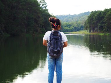 Gezgin konseptinde gölün ve dağın önünde duran Asyalı kadın sırt çantası vadi ve nehir altyapısı olan çanta taşıyor.