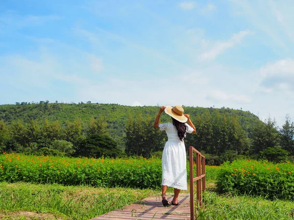 Asian girl holding the hat  travel in flower parkland with blue sky and mountain.Woman traveler in white dress standing in flower park with meadow and valley.