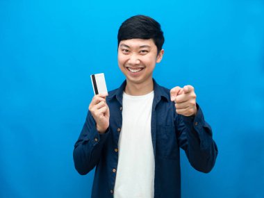 Neşeli adam gülümsüyor, elinde kredi kartı ve işaret parmağıyla mavi arka planını gösteriyor.