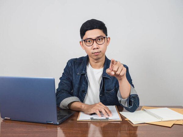 Young businessman wear glasses sit at workspace point finger at you