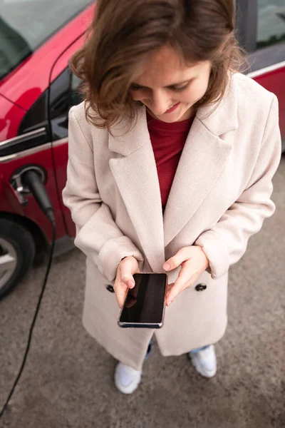 赤い電気自動車の横にスマートフォンでベージュコートの女性 — ストック写真