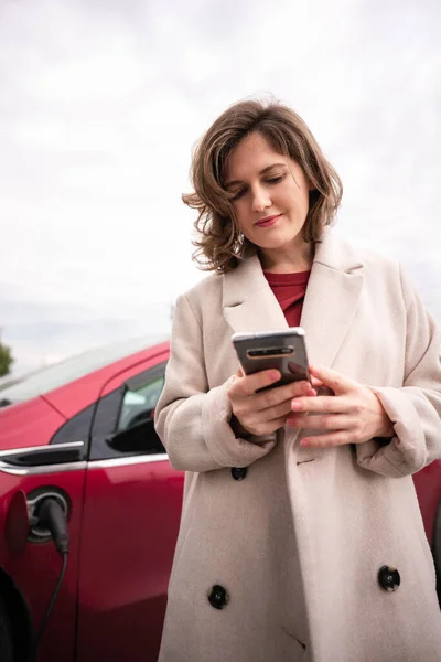 Vrouw in beige jas met smartphone naast elektrische auto — Stockfoto