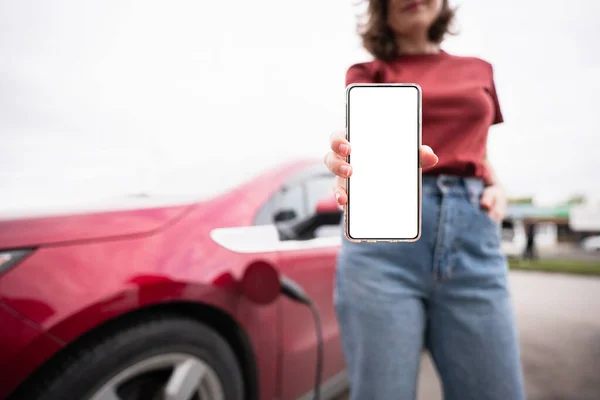 Een vrouw op de achtergrond van een rode elektrische auto toont een lege smartphone scherm — Stockfoto