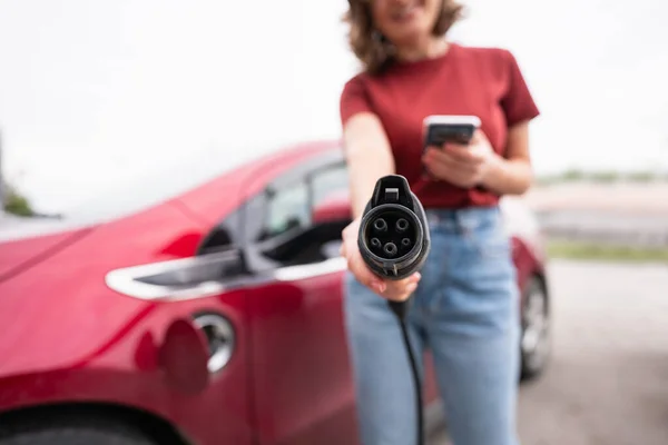 Vrouw met smartphone toont elektrische auto opladen plug — Stockfoto