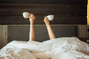 Yataktaki battaniyenin üzerinde bir çift el kahve bardaklarını tutuyor. Sabah oldu.