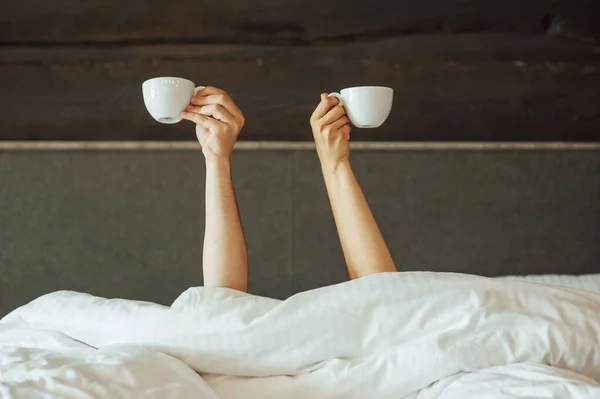 O pereche de mâini se țin deasupra păturii din pat țin cești de cafea. Dimineața — Fotografie, imagine de stoc