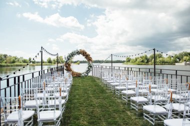 Nehir kenarında bir düğün merasimi için yuvarlak kemer. Beyaz cam sandalyeler misafirler içindir.