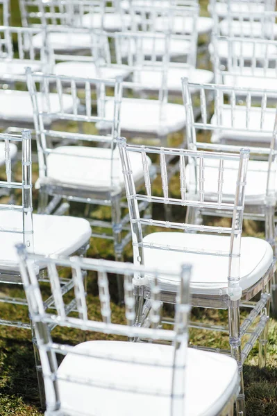 Οι λευκές γυάλινες καρέκλες στέκονται στο πράσινο γρασίδι. Στρογγυλή αψίδα για μια γαμήλια τελετή στο βάθος — Φωτογραφία Αρχείου