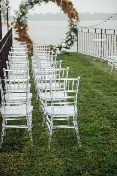 Bílá skleněná křesla stojí na zelené trávě. Kulatý oblouk pro svatební obřad v pozadí. Deštivý den — Stock fotografie