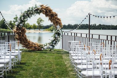 Nehir kenarında bir düğün merasimi için yuvarlak kemer. Beyaz cam sandalyeler misafirler içindir.