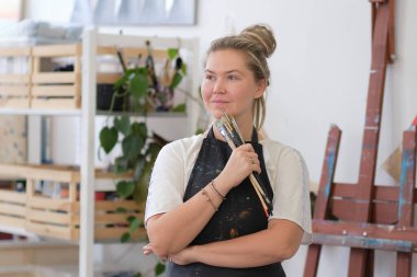 Kapalı alanda fırçayla poz veren yetenekli bir kadın sanatçı. Fırça tutarken kollarını kavuşturur, gülümseyerek kameraya bakar. Geniş tuvalli otantik yaratıcı stüdyo.