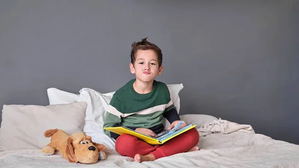 Αστείο, γοητευτικό αγοράκι που διαβάζει αλφάβητο. Το παιδί κοιτάζει το ανοιχτό βιβλίο. Μάθημα κατ 'οίκον. Συμπυκνωμένο αγοράκι που κάθεται στο κρεβάτι διαβάζοντας βιβλίο στο σπίτι — Φωτογραφία Αρχείου