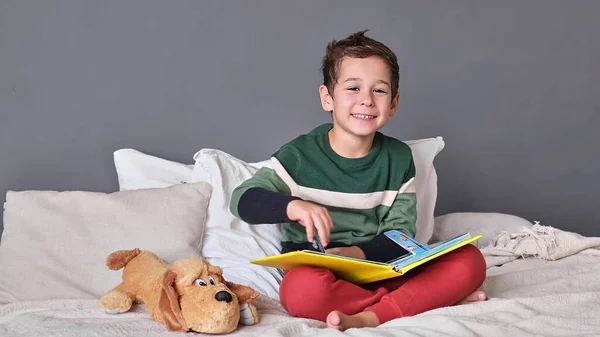 Divertente, affascinante bambino che legge alfabeto libro. Il bambino guarda il libro aperto. Lezione di scuola a casa. Ragazzino concentrato seduto sul letto lettura libro a casa — Foto Stock