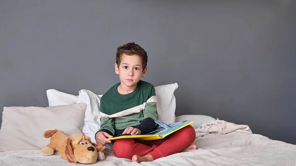 Χαριτωμένο αγοράκι που διαβάζει αλφάβητο. Το παιδί κοιτάζει το ανοιχτό βιβλίο. Μάθημα κατ 'οίκον. Συμπυκνωμένο αγοράκι που κάθεται στο κρεβάτι διαβάζοντας βιβλίο στο σπίτι — Φωτογραφία Αρχείου