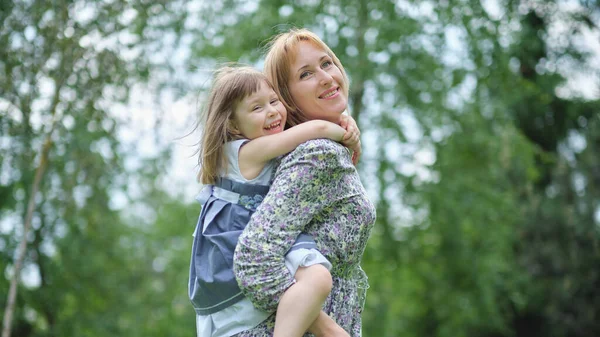 Family having fun outdoors. Little adorable daughter piggybacks her mother. Mom carrying loving kid on her back laughing enjoy funny time together. — Stock Photo, Image
