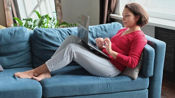 İnternetten alışveriş yapan orta yaşlı bir kadın. Kadın evde dizüstü bilgisayar kullanıyor. Tahta koltukta bir haftasonu. Not defteri ekran tipi iletiye bak. bilgisayar interneti — Stok fotoğraf
