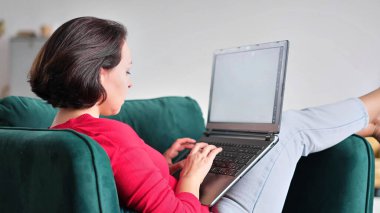 Orta yaşlı bir kadın internetten alışveriş yapıyor. Kadın evde dizüstü bilgisayar kullanıyor. Tahta koltukta bir haftasonu. Not defteri ekran tipi iletiye bak. bilgisayar interneti