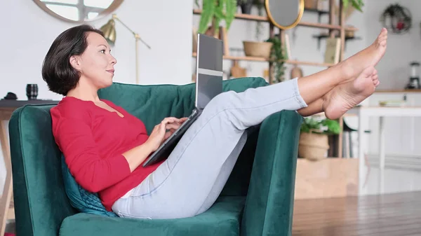 Középkorú nő online vásárol. A nő laptopot használ otthon. hétvége fából készült fotelban. notebook nézd meg a képernyő típusú üzenetet. számítógép internet — Stock Fotó