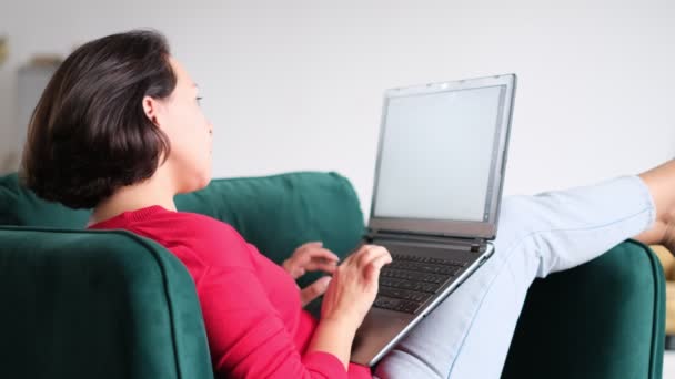 Orta yaşlı bir kadın internetten alışveriş yapıyor. Kadın evde dizüstü bilgisayar kullanıyor. Tahta koltukta bir haftasonu. Not defteri ekran tipi iletiye bak. bilgisayar interneti — Stok video