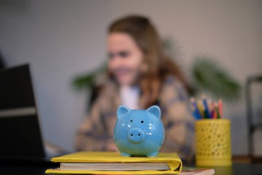 Mavi domuz kumbarası ve ders kitapları masanın üstünde, arka planda genç bir kız dizüstü bilgisayarda ödev yapıyor. eğitim maliyeti kavramı