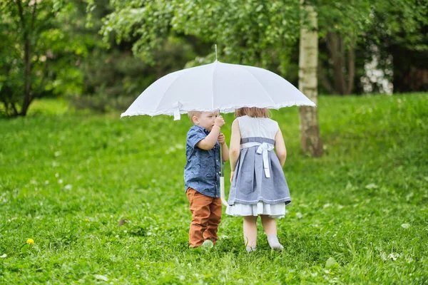 Rapariga e rapaz estão escondidos debaixo de um guarda-chuva. irmão e irmã estão brincando no parque. — Fotografia de Stock